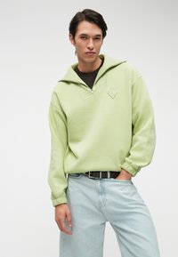 Ανοιχτό πράσινο φούτερ με φερμουάρ και γιακά, με διαχρονικό διαμαντένιο λογότυπο. Συνδυάζεται με ανοιχτό μπλε τζιν σε χαλαρή γραμμή και μαύρη ζώνη.