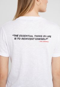 Žena v bílém tričku s citátem "Základní věc v životě je znovu se vynalézat" od Karla Lagerfelda, napsaným černým a červeným písmem.