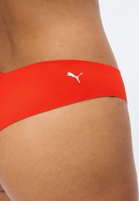 Közeli kép egy személyről, aki piros Puma fürdőruha alsót visel, oldalról látható a csípő és a Puma-logó a anyagon.