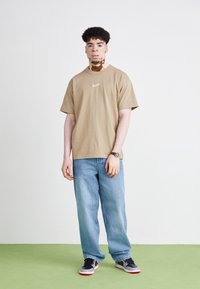 Beige Nike-T-Shirt mit kleinem Logo, kombiniert mit weiten hellblauen Jeans und schwarzen Sneakers mit roten Akzenten, stehend auf grünem Boden.