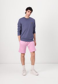 Sweatshirt gris-violet avec un logo jaune, associé à un short rose. Le mannequin porte des baskets blanches et des chaussettes de cheville blanches.