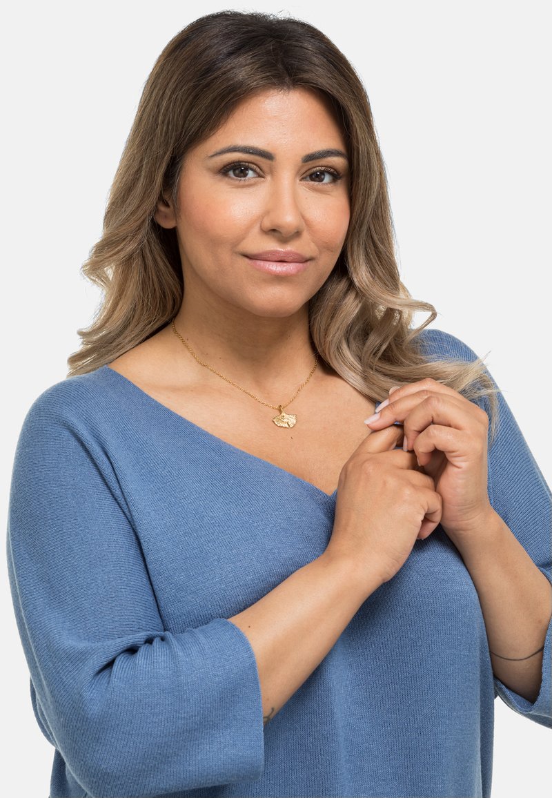 Hellblauer gestrickter Pullover mit weiter V-Ausschnitt, Dreiviertelärmeln, getragen von einer Person, die den Stoff hält. Goldene Halskette mit Muschel-Anhänger sichtbar.