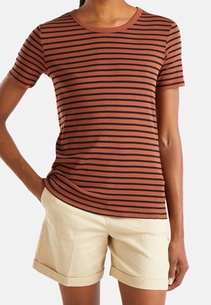 Kvinde iført en rustfarvet og sort stribet kortærmet skjorte sammen med beige shorts med opbuk ved kanten, stående med den ene hånd i lommen.