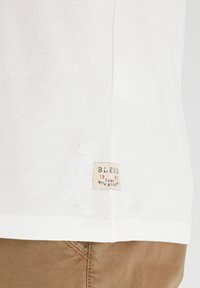 Hvit bomulls T-skjorte med et sydd merke på siden. Merket har "BLEND," året 1993, og teksten "WITH ATTITUDE."