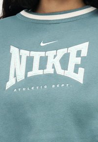 Πουλόβερ σε τυρκουάζ με λευκή ριμπ λαιμόκοψη που φέρει τη λέξη "NIKE", μικρό λογότυπο swoosh και το κείμενο "ATHLETIC DEPT."