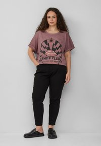 Lila T-Shirt mit kurzen Ärmeln und schwarzem Grafikdesign, das ein Sonnen- und Bergmuster zeigt, kombiniert mit schwarzen Hosen und Slippern.