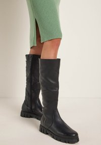 Schwarze knielange Lederstiefel mit einer strukturierten Oberfläche, seitlichen Reißverschlüssen und dicken Gummisohlen, kombiniert mit einem grünen gerippten Kleid.