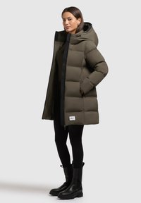 Pufová bunda v olivově zelené barvě s černým zipem a kapucí. Má prošívaný design s cedulkou na dolním předním dílu. Nosená přes černé legíny.