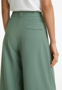 Pantalons larges à carreaux vichy verts, taille haute, avec une poche plate au dos et une texture lisse ; présentés de côté avec une main reposant sur la hanche.