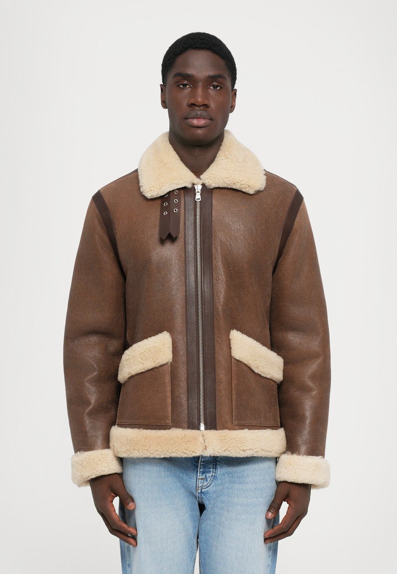 Mann trägt eine braune Lederjacke mit einem cremefarbenen Shearling-Kragen, -ärmeln und -taschen, kombiniert mit hellblauen Jeans, und steht vor einem weißen Hintergrund.
