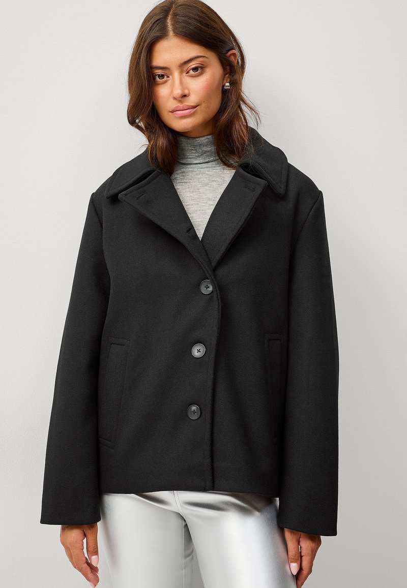 Cappotto nero in misto lana con colletto a punta, tre bottoni e tasche laterali. Presenta una vestibilità comoda e un dolcevita grigio chiaro a contrasto sotto.