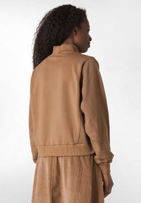 Giacca in morbido tessuto marrone con colletto alto e polsini a coste, caratterizzata da una vestibilità rilassata e una texture liscia. Vista da dietro.