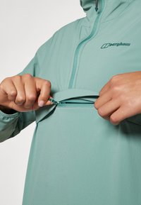 Mintvihreä kangaspullover, jossa on edessä vetoketju ja taskuaukko, piilohuppu naruilla ja joustavat hihansuut. Brändilogo näkyvissä.