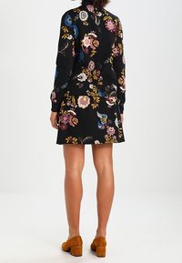 Svart klänning med blommönster och långa ärmar, som visar rosa, blå och gula blommor; matchad med bruna högklackade skor.