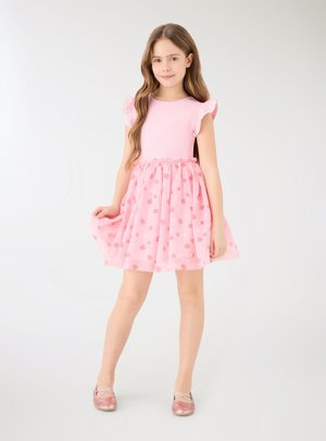 Ragazza giovane con lunghi capelli castani che indossa un vestito rosa con maniche arricciate e a pois, in piedi e che tiene la gonna con una mano.