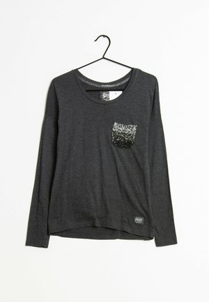 Camisa gris de manga larga con cuello redondo y un bolsillo adornado con lentejuelas negras y plateadas. La tela es suave y ligeramente texturizada.
