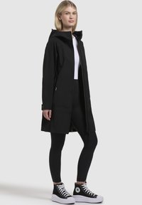 Black waterproof jacket with a hood, featuring a straight cut, long sleeves, and a zip front. Worn over black leggings and high-top sneakers.