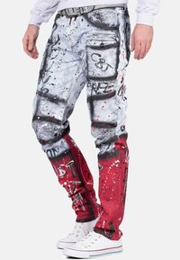 Pantaloni di denim multicolore con un design a macchie in grigio e rosso. Inclusi più tasche e elementi testuali grafici.