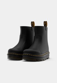 Botines negros de tobillo hechos de un material sintético suave, con una suela gruesa, costuras amarillas y lengüetas para un fácil calzado.