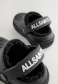 Zwarte rubberen klompen met een zachte zwarte bontvoering, voorzien van een brede band met "ALLSAINTS" in witte letters over de bovenkant.