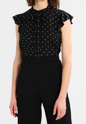 Vrouw draagt een zwarte mouwloze blouse met witte polka dots en geplooide schouders, gecombineerd met zwarte high-waisted broek.