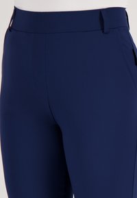 Close-up van marineblauwe fitted broek met riemlussen en zakken aan de voorkant, gedragen op een witte achtergrond.