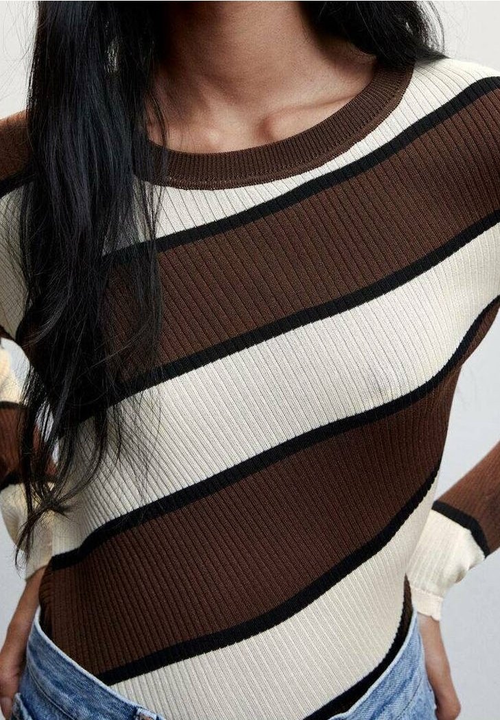 Nainen, joka pukeutuu resoroituun neulepaitaan, jossa on vinoja ruskeita, kermanvärisiä ja mustia raitoja, yhdistettynä sinisiin farkkuihin.