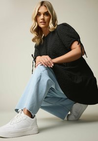 Top oversize nero texture con maniche corte, abbinato a jeans a gamba larga blu chiaro e sneakers bianche. Dettagli di nodo notevoli sulle maniche.