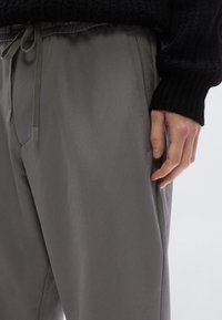 A világosszürke nadrág rugalmas derékpánttal és húzózsinórral, ferdén zsebekkel és sima textúrával rendelkezik, fekete kötött pulóver mellett bemutatva.