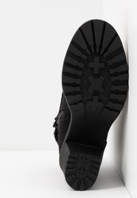 Semelle de botte noire avec un motif de semelle profond en formes de croix, inclinée contre une surface blanche à angles.