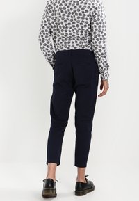 Henkilön takakuva, jolla on valkoinen paita mustilla lehtikuvioilla ja tummansiniset kapenevat housut, yhdistettynä mustiin kenkiin ja näkyviin takataskuihin.