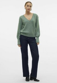 Maglione verde lavorato a maglia con scollatura a V, caratterizzato da un motivo a zigzag testurizzato, abbinato a jeans blu scuro a gamba dritta e stivaletti neri alla caviglia.