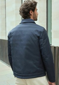 Jachetă călduroasă, de culoare albastru marin, cu un design cu guler, având o textură netedă și un aspect minimal, văzută din spate.