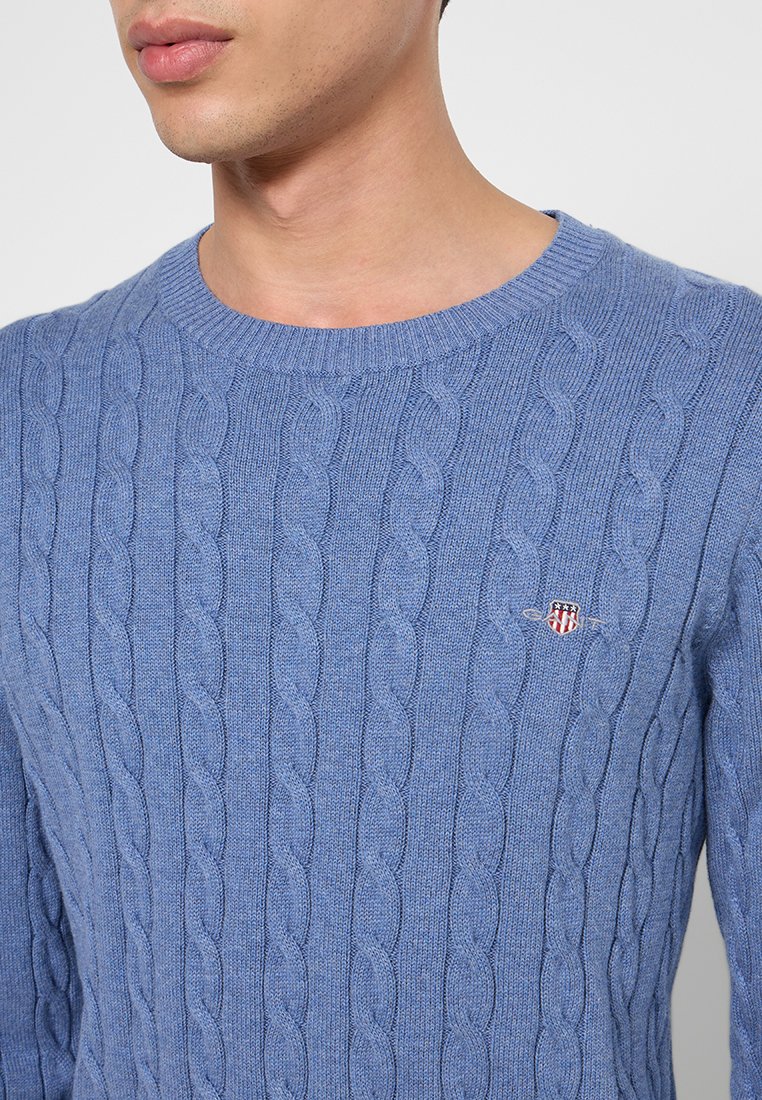 Suéter de punto azul claro con un patrón de trenzas, escote redondo y un pequeño emblema en el pecho. Textura suave y diseño ajustado.