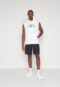 Biely bezrukávový mikina s logom "Champion", v kombinácii s tmavými šortkami. Model má na sebe biele tenisky a ponožky s logom. Jednoduchý, moderný dizajn.