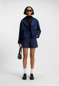 Veste courte bleu marine avec de grands boutons, portée sur un haut en tricot noir et un short assorti. Le mannequin tient un petit sac noir et porte des lunettes de soleil.