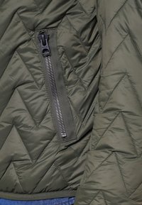 Tmavě zelená prošívaná bunda s šikmým prošíváním a vertikální černou zipovou kapsou na přední levé straně.