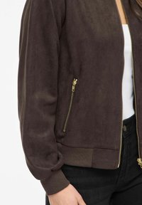 Brūna zamša bumbvedēja jaka ar rāvējslēdzēja priekšpusi, ribveida manšetēm un divām sānu kabatām ar rāvējslēdzējiem. Gluda tekstūra un ikdienišķs dizains.