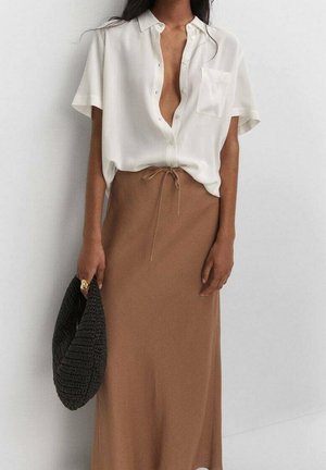 Kobieta w luźnej, białej, krótkoobcisłej bluzce z guzikami i brązowej spódnicy o wysokim stanie, trzymająca czarną, teksturowaną torebkę przy białej ścianie.