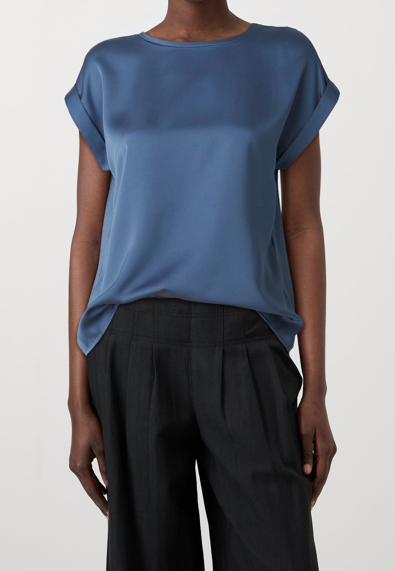 Femme portant un chemisier bleu ample à manches courtes, rentré dans un pantalon noir taille haute à jambes larges, sur un fond uni.