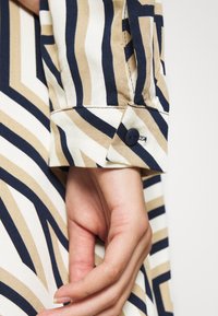 Main reposant près du poignet avec une chemise à manches longues aux rayures beige, marine et blanche, boutonnée avec un bouton foncé.