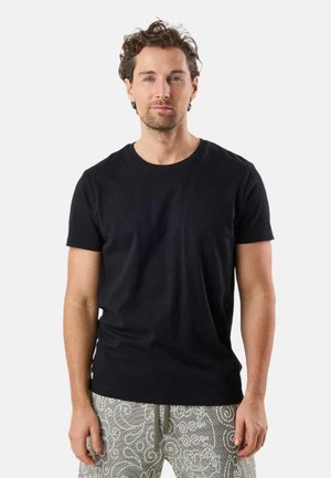 Mand med krøllet hår iført en ensfarvet sort t-shirt og grå mønstrede shorts, stående foran en hvid baggrund.