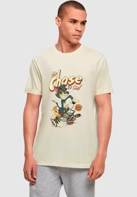 Beige bomulds t-shirt med et tegneserieprint af en blå kat og en brun mus i en racerbils, med farverig tekst der siger "Jagten er i gang!"