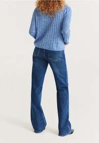 Kiharatukkainen henkilö, joka pukeutunut vaaleansiniseen ribbineulottuun neuleeseen ja tummansinisiin leveälahkeisiin farkkuihin, seisoo selin kädet taskuissa.