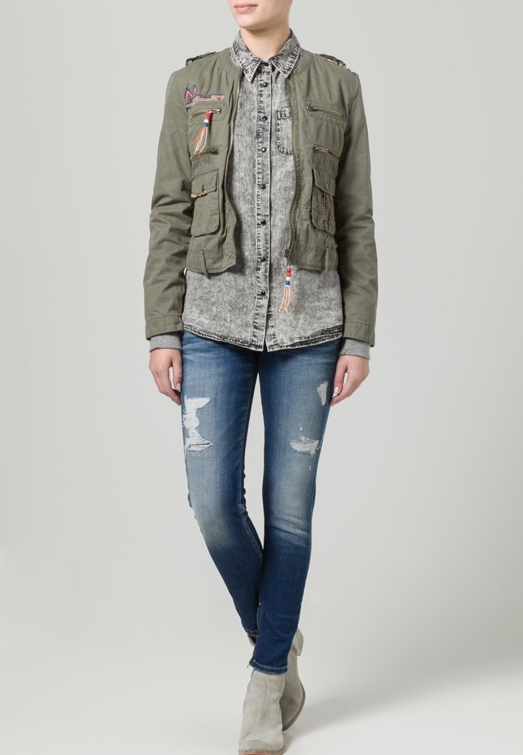 Cazadora bomber color verde oliva con múltiples bolsillos, camisa gris con estampado debajo y jeans azules desgastados, combinados con botas gris claro.