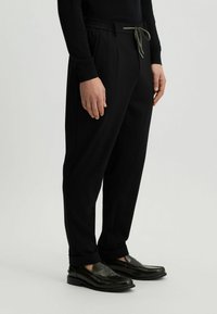 Schwarze, tapered Hosen mit elastischem Bund und Kordelzug. Glattes Material, seitliche Taschen, kombiniert mit dunklen Slippern.