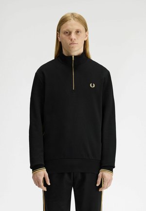 Giovane con capelli lunghi che indossa una felpa nera con zip corta e pantaloni coordinati con polsini e strisce laterali bordati d'oro.