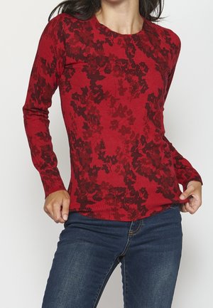 Femme portant un pull rouge à manches longues avec un motif floral sombre et un jean bleu, tenant les bords inférieurs du pull.