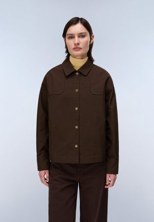 Kvinde med kort mørkt hår iført en mørkebrun knappet jakke over en gul rullekravesweater stående mod en lyse baggrund.