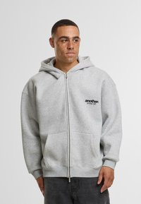 Gray zip-up hoodie made of soft material, featuring a front pocket, ribbed cuffs, and a black logo on the chest. Simple, casual design.
